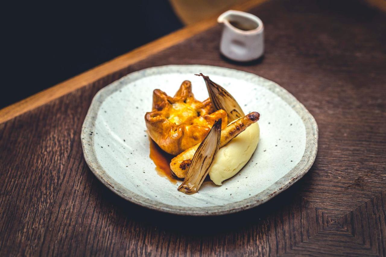 The Grand, York Hotel Kültér fotó The photo shows a beautifully presented plate of food. At the center, there is a savory dish that appears to be a type of dumpling or bun, topped with a golden sauce. Surrounding it are two elongated, charred elements, possibly grilled vegetables or 